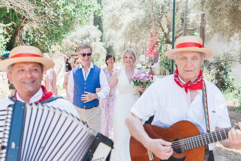 Bride and her father walking to the ceremony while being serenaded by a trio of Greek musicians