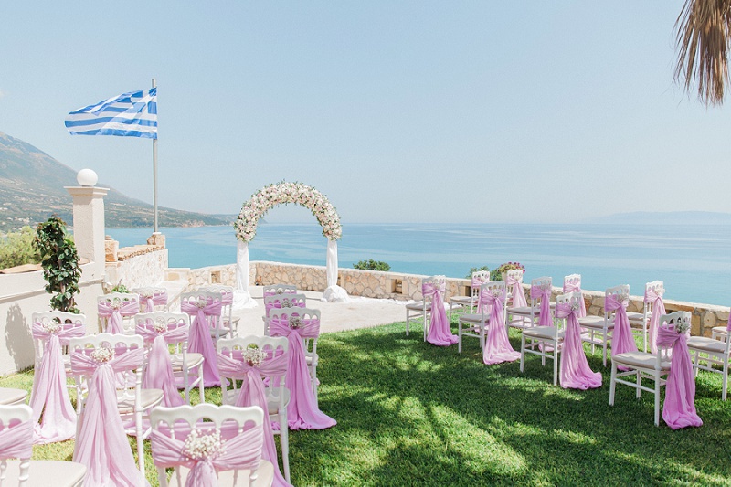 The Villa Rosa ceremony area with Greek flag, floral arch and decorated white chairs.