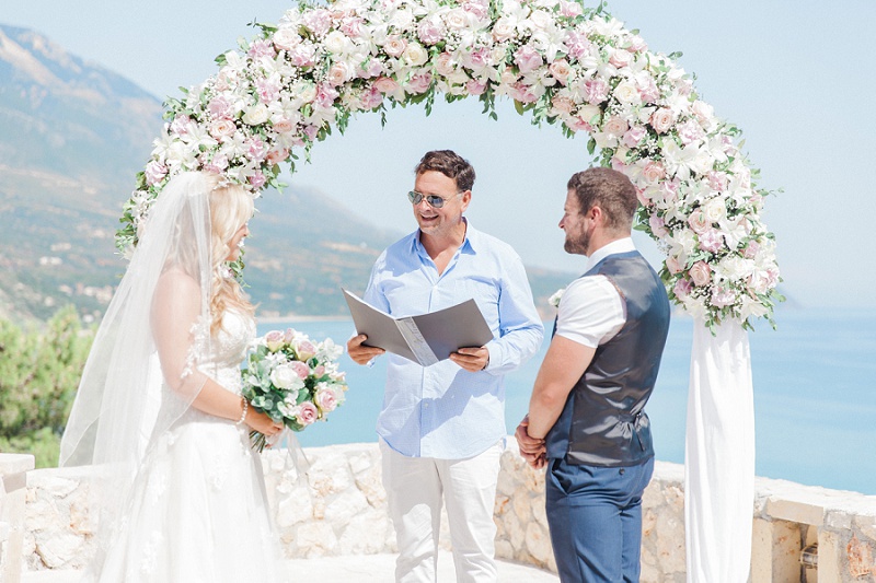 Bride and Groom exchanging vows under a pink and white floral arch with a view of the sea.