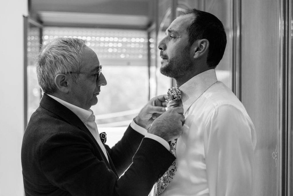 The groom is helped with his tie by a friend on the morning of his wedding