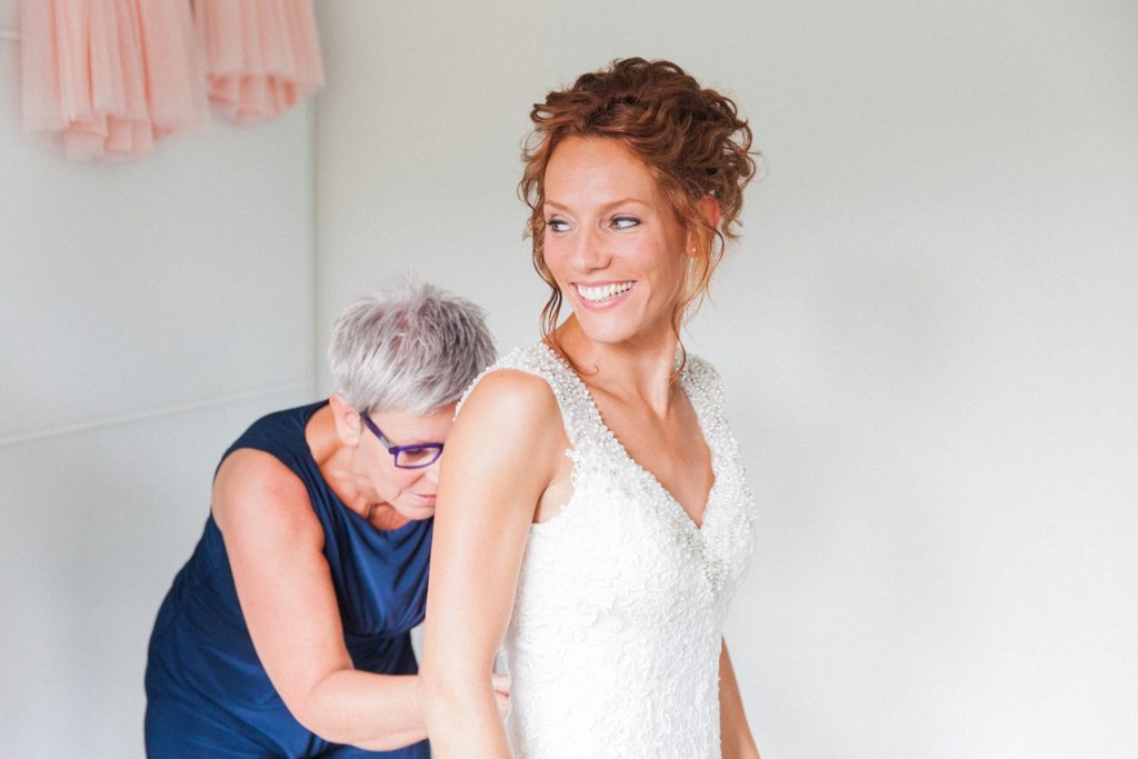 Bride smiles as her mother helps her put her Justin Alexander wedding gown on.