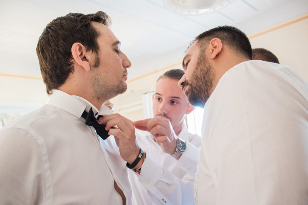 Groomsmen help the groom put on his bowtie on the morning of his wedding in Lefkada