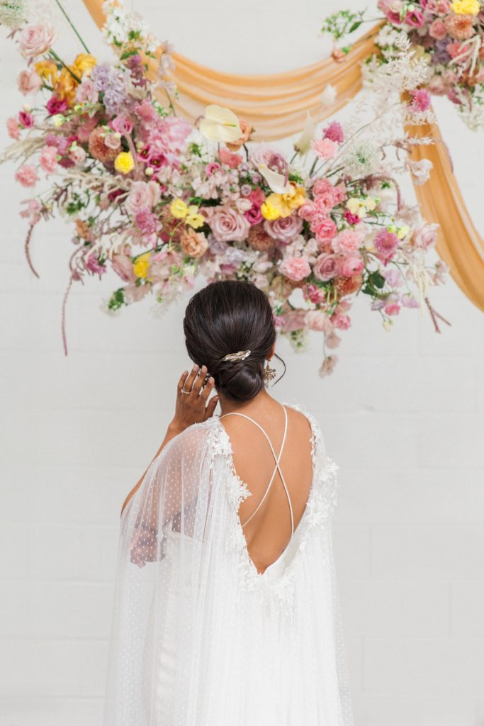 Bride stands under colourful hanging floral arrangements in a Halfpenny London wedding dress