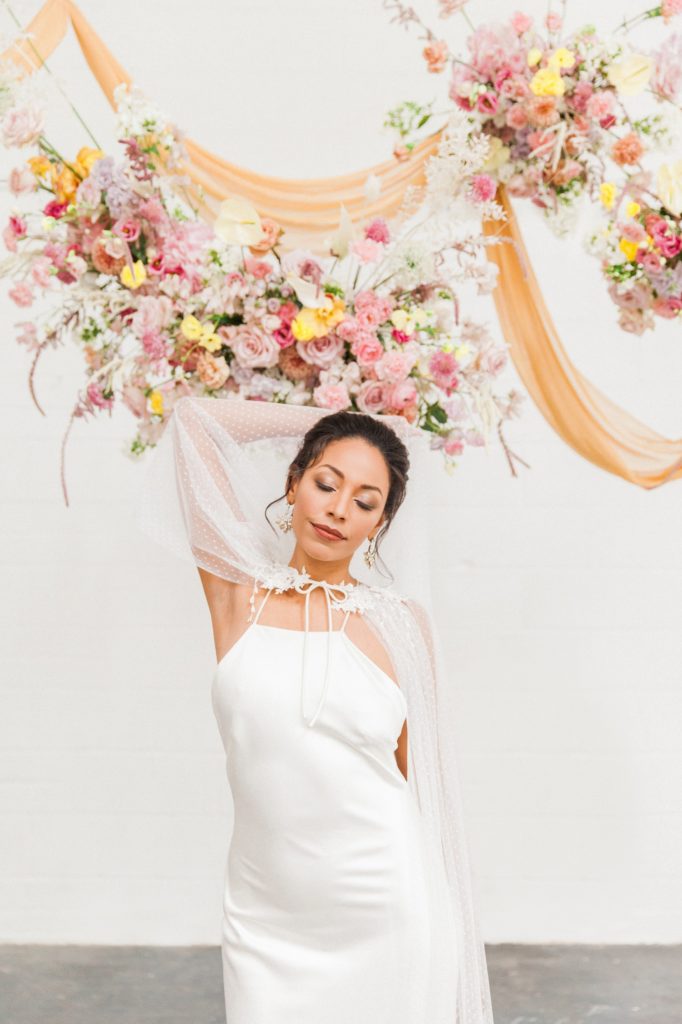Portrait of a black bride in a Halfpenny London wedding dress below colourful hanging flower arrangements