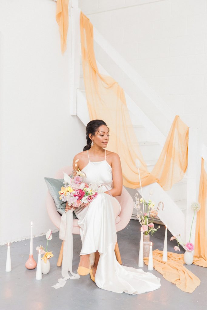 Beautiful black bride sitting in a pink lounge chair surrounded by colourful flowers and candles