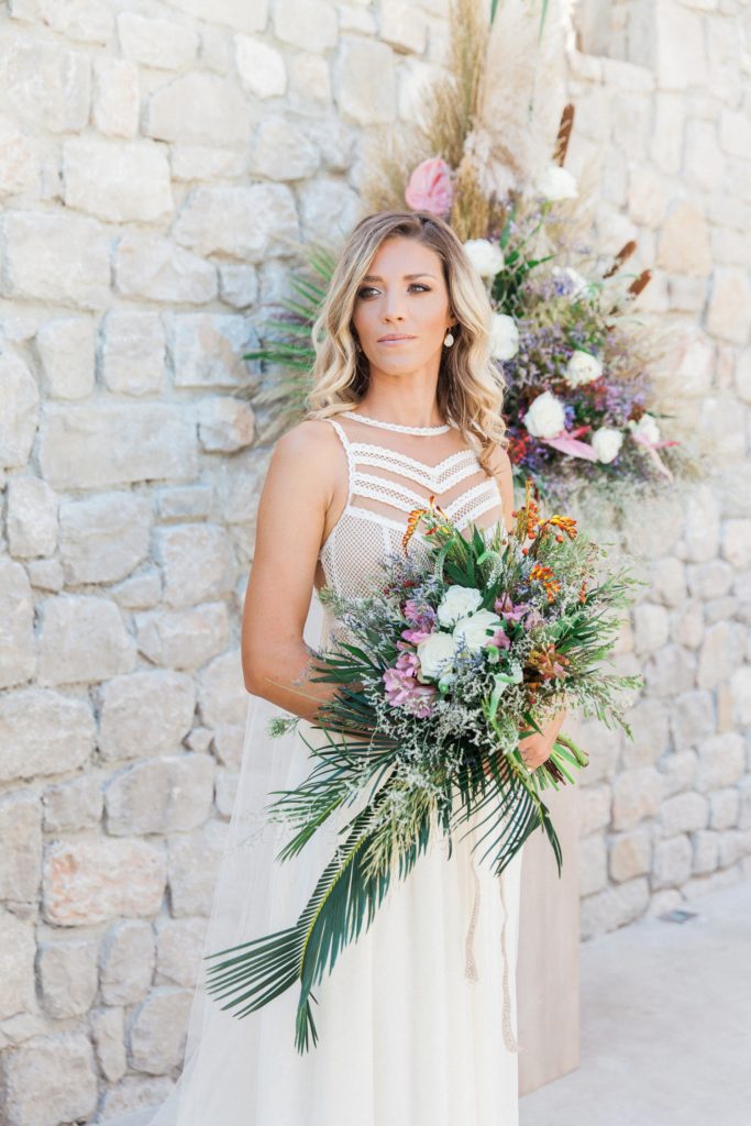 Boho bride poses with her bouquet in her wedding ceremony area at Crystal Waters Lefkada