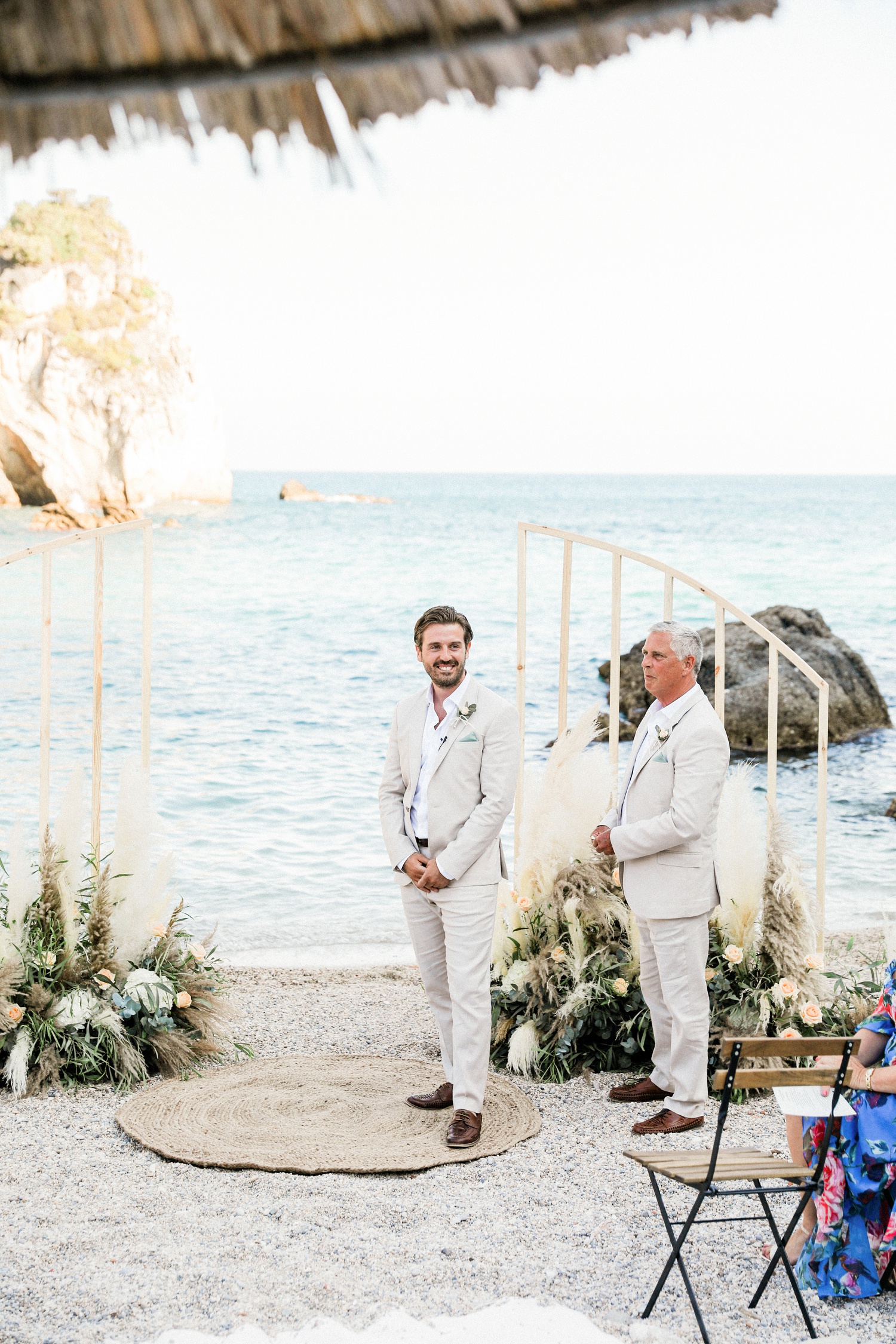 Groom standing in front of a boho wedding arch smiles as he sees his bride arrive