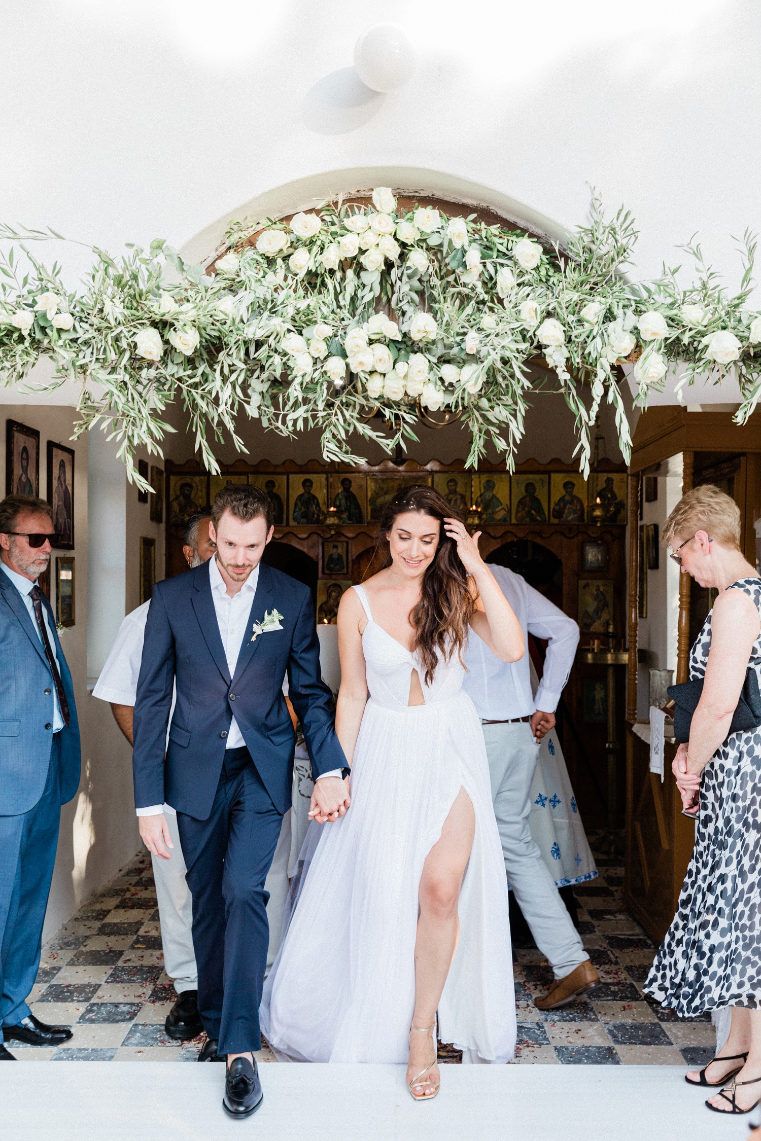 Bride and groom walk out of the orthodox church under a garland of olive and white roses