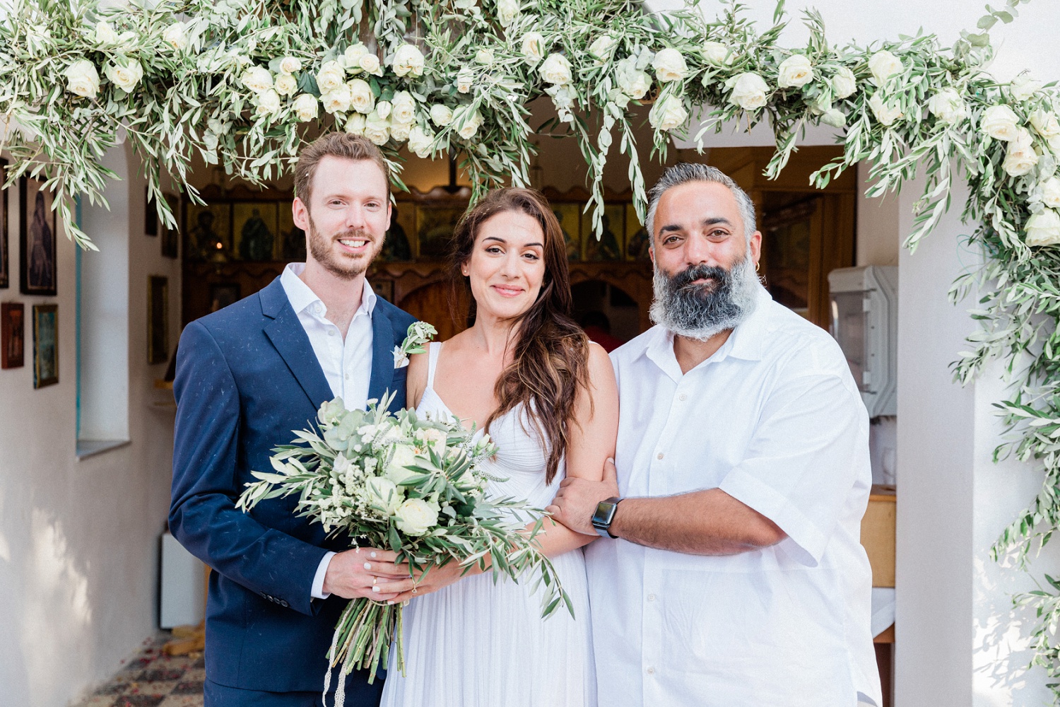 Couple stand with their best man under a garland of olive and white roses