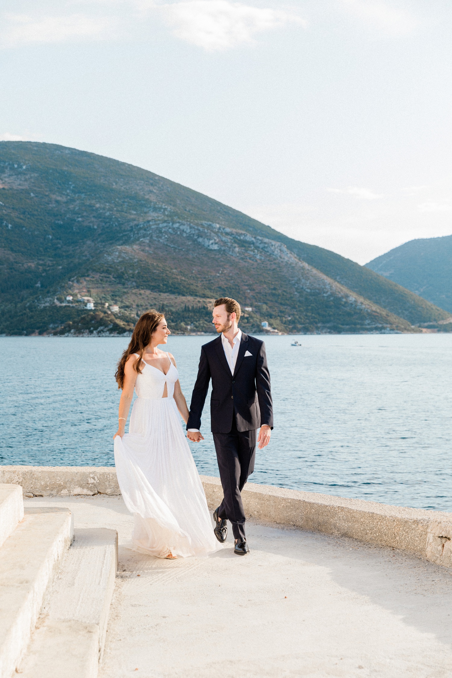 Bride and groom walk together on a terrace above the sea with Ithaca behind them