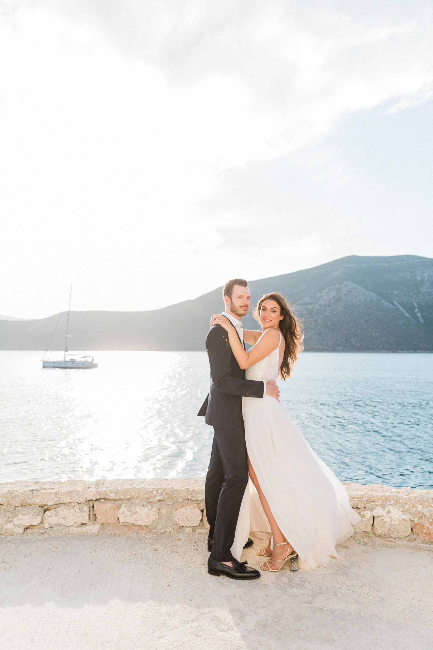 Bride and groom stand together at sunset on a terrace against a view of Ithaca and a sailing yacht