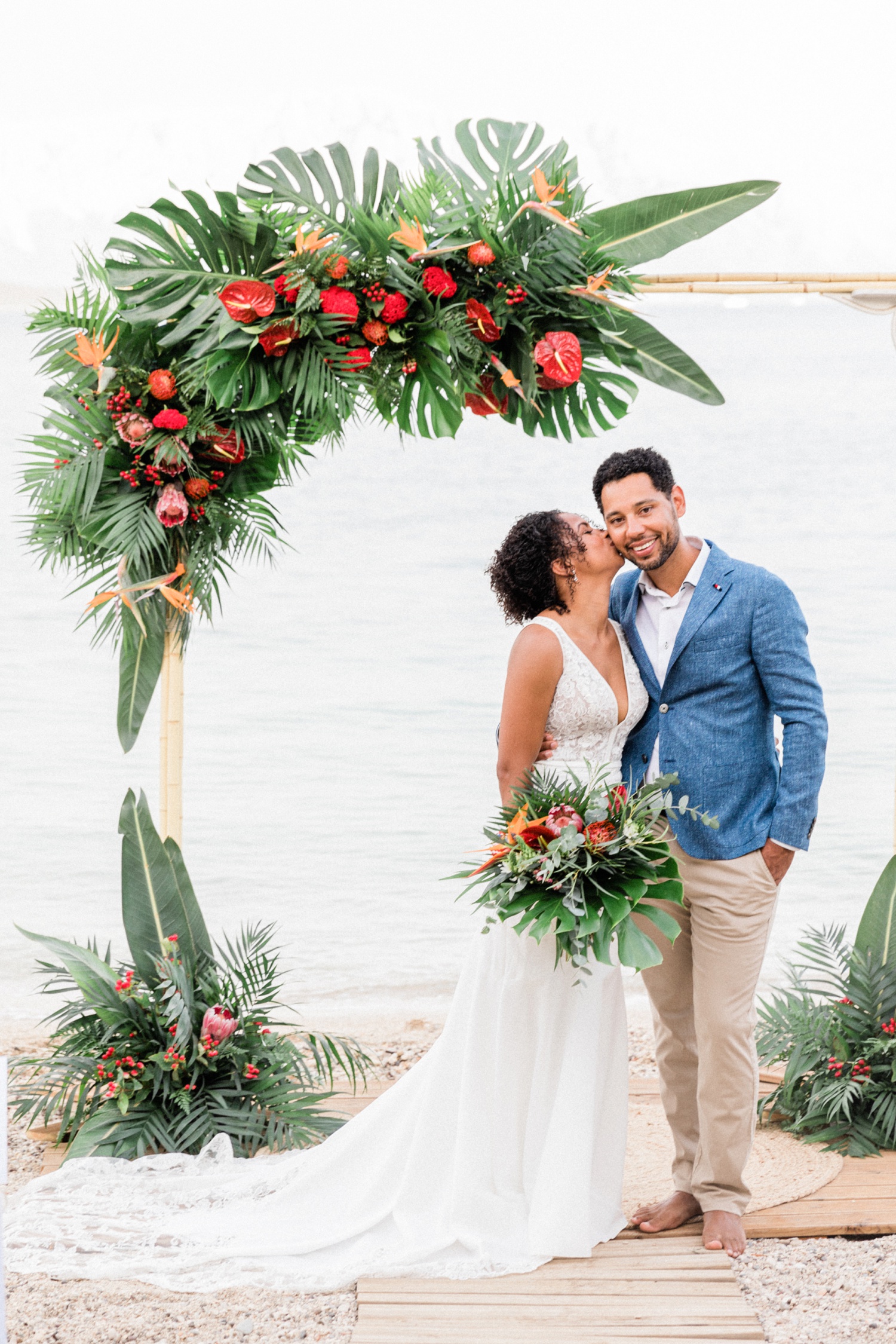 Bride kisses groom on the cheek under a tropical wedding arch on the beach